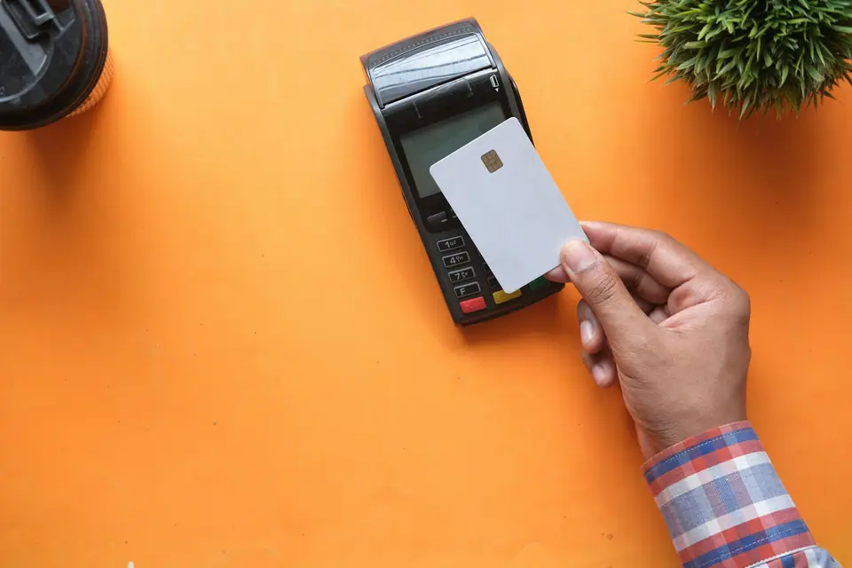 Hand holding a plain blank payment card against a card terminal on an orange surface